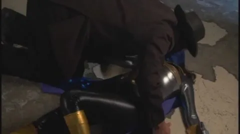 Asian woman in gold armor touched by men in black suits